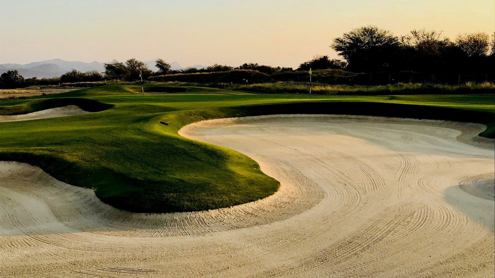A large raked bunker curving around the edge of a green at dusk, with two flags and distant hills behind