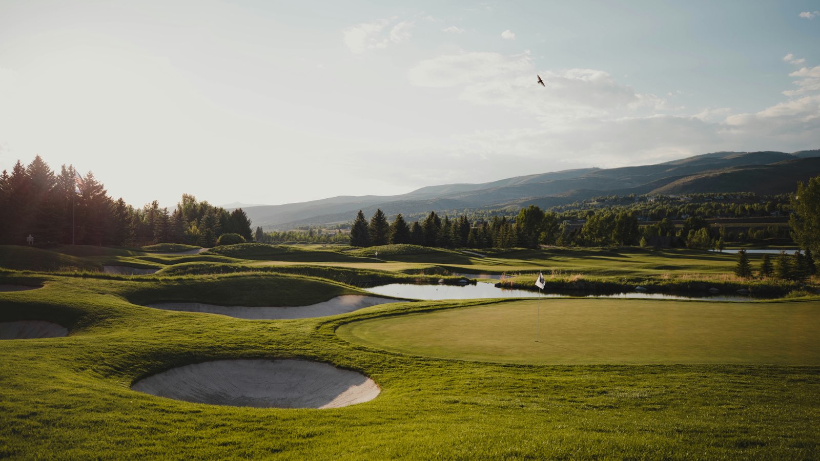 A golf green with the flag in, framed by bunkers and a pond, with wooded hills and mountains behind