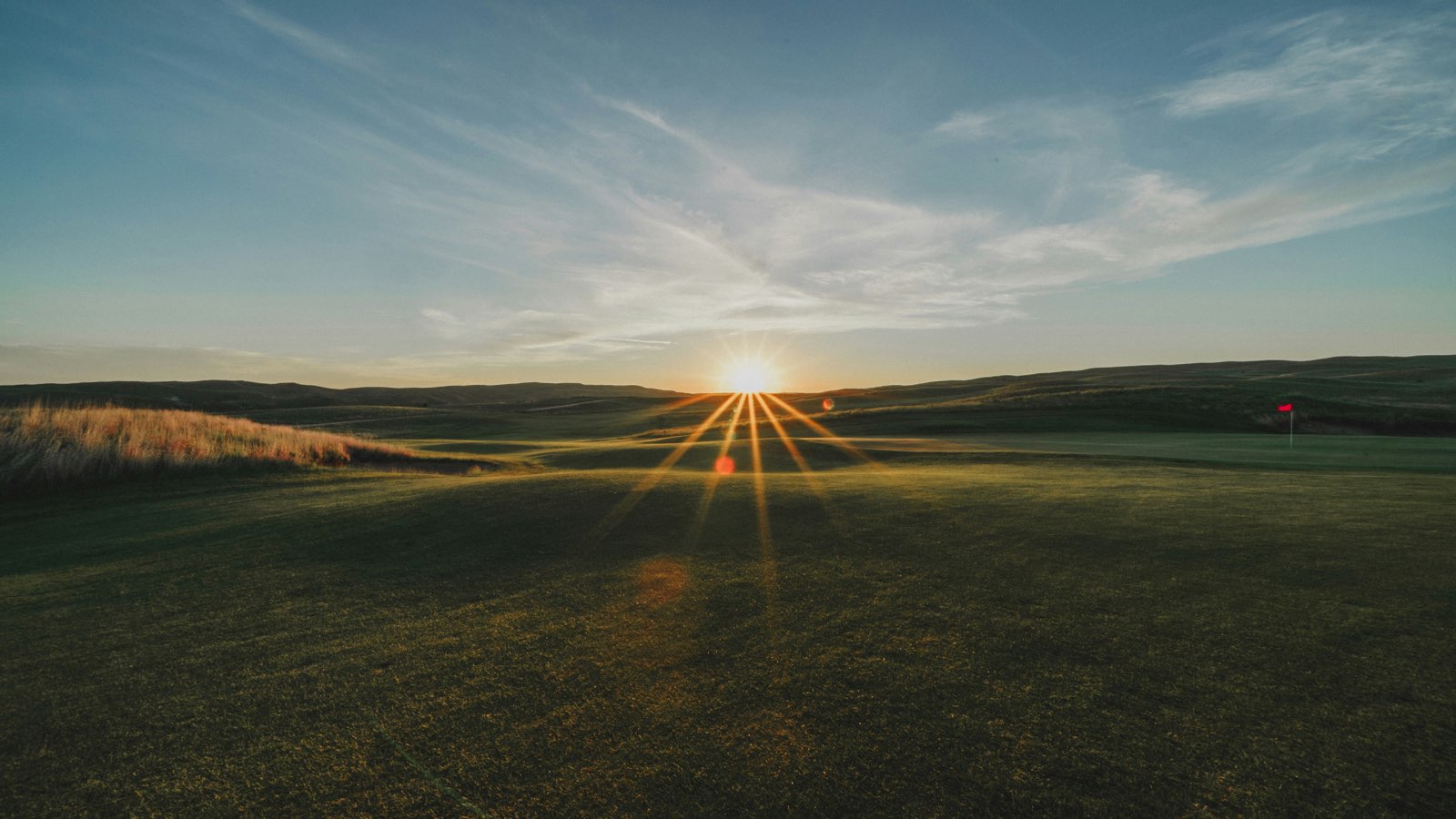 A golf fairway at sunset with the sun starbursting low over distant hills and a red flag on the green in the distance
